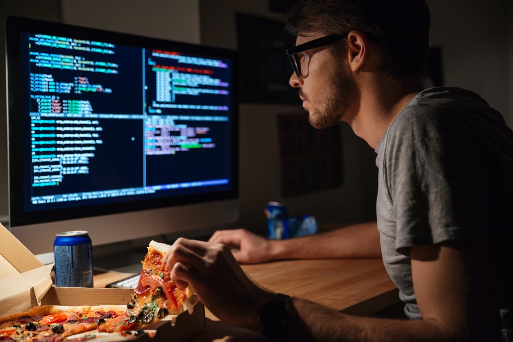 Homem está digitando de frente ao computador. enquanto pega uma pizza.