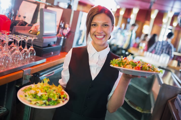 Mulher uniformizada carregando em cada mão um prato com bastante salada.