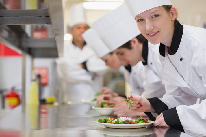 Alunos de culinária uniformizados estão exibindo seus pratos numa bancada.